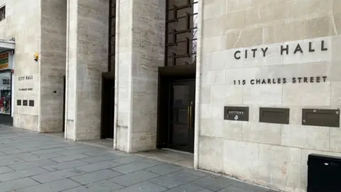 The exterior of the entrance to City Hall in Leicester, the City Council's headquarters. It has a light stone wall and three arches with black doors.