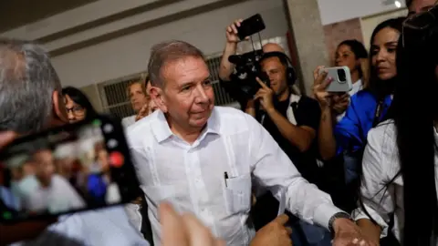 Reuters: Edmundo González passa por várias pessoas segurando câmeras. Ele tem cabelo castanho curto e veste uma camisa branca com uma caneta saindo do bolso da camisa.