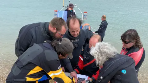 A group of people wearing scuba diving gear are gathered in a circle and looking at and pointing down to an image on what looks like paper. The shoreline and the top of a boat are in the background.