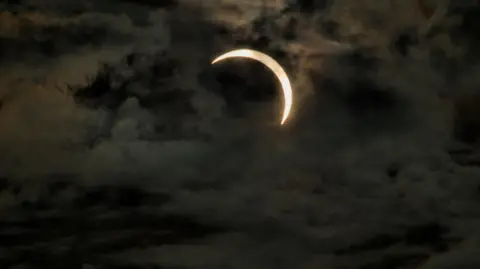 BBC Weather Watchers/Phil Powe A partial solar eclipse captured in Whitehaven. A thin sliver of sun is visible behind think dark clouds.