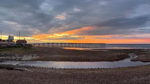 BBC Weather Watchers/Francis Annett Saltburn beach which is covered in pebbles. A large pool of water is flowing into the sea through the middle of the beach. A pier can be seen in the distance. The sky is grey and orange.