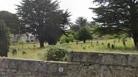 Google A cemetery with dozens of gravestones. Grass surrounding them has not been cut and is overgrown