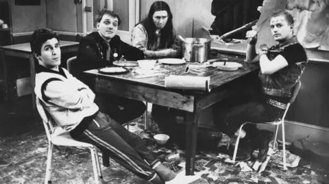 Don Smith/Radio Times/Getty Images A black and white photo of four white males - Christopher Ryan, Rik Mayall, Nigel Planner and Adrian Edmondson - sitting around a table in a scene from the television sitcom 'The Young Ones'
