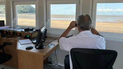 NCI Minehead A man in uniform is sitting in a chair looking out past the coastline using binoculars.