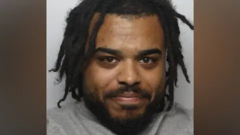 BBC A police mugshot of a man wearing a grey top, standing against a light-coloured background. He has black hair and dreadlocks.