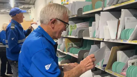 The image shows a man with white hair and spectacles sorting letters on a shelf, wearing a blue polo shirt. In the background are other workers with blue jackets and caps on, who are also sorting letters.