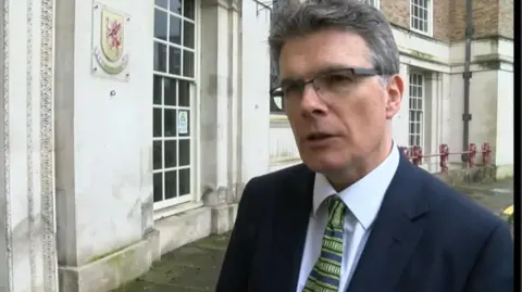 Julian Wooster with short grey hair and glasses wearing a dark blue jacket, white shirt and green tie, standing outside a stone-built council building