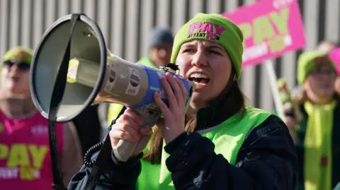 A female union member speaking animatedly into a megaphone. She has long brown hair and wears a lime green hat with the slogan "pay attention". 