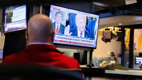 Stock market trader in red jacket faces a TV screen showing Trump being sworn in as US president 