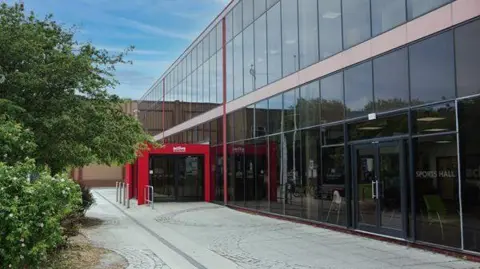 Northumberland County Council The entrance to Concordia Leisure Centre, a two-storey building clad in tinted glass from floor to roof. The steel work and entrance area is red. There are trees to the left of a block paved pathway.