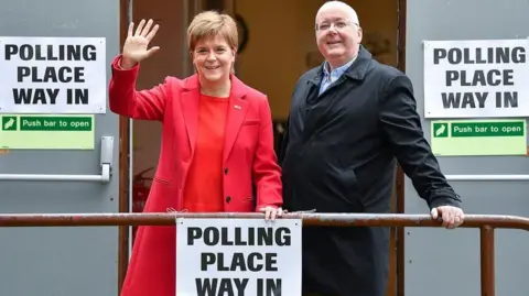 Nicola Sturgeon in a red coat and Peter Murrell in a black coat standing outside a polling place. They are smiling and she is waving.