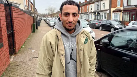 A man wearing a grey hoodie and beige jacket stands on the pavement, looking into the camera. Cars can be seen parked on the road behind him.