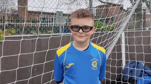 Leo a young boy with a blue and yellow football shirt on and stood in a football goal with his hands behind his back. He is looking at the camera and wearing black rimmed glasses with tinted lenses.
