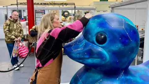 A woman in a brown apron is painting the head of a 1.5m high duck statue that has been decoratively painted in a marketplace entrance. The woman has long blonde hair, a pink and black jumper, black leggings and boots. The duck is painted in blue and green swirls with white constellations and stars also painted on. A man in a camouflage jacket, jeans and holding two shopping bags is looking at the duck from behind the black security rope.