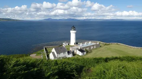 Lee Cassidy A lighthouse and outhouses on the grassy edge of the coast. Trees are seen in the foreground and beyond the coast is the deep blue sea.