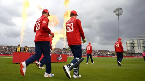 England players, in their T20 kit of dark blue trousers and red tops, walk out at the County Ground in Bristol as flames shoot up as part of a pyrotechnic display