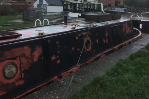 Warwickshire Police A narrowboat with circular windows painted black with patchy red paint. The craft sits next to a lock and alongside the concrete edge of a towpath. Behind it are several buildings.