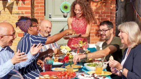 Eden Project The picture shows a group of adults gathered around a table outdoors, sharing food together in what looks like a neighbourhood setting. They are seated close together around a bright blue table filled with plates and bowls of food, including strawberries, cakes, salads and drinks. Several hands are reaching across the table to pass plates and serve food.