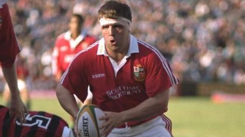 Tom Smith with dark hair in a headband playing rugby