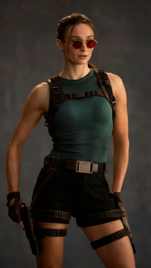 Amazon MGM Studios A photo shows Sophie Turner as Lara Croft. She wears red-tinted sunglasses, her hair tied back. She wears a green vest top and tight, thigh-length shorts. One of her fingerless-gloved hands rests on a pistol in a thigh holster.