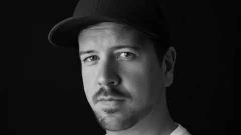 A black and white image of Russell Bowen, who is looking solemnly right at the camera. He is wearing a black cap and white t-shirt. He has short dark hair and facial hair.