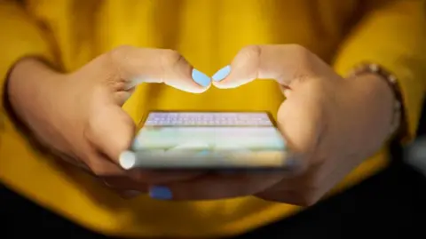 Close-up view of an adult's hands holding a smartphone. The person, who cannot be identified in the picture, has a yellow top and light blue nail polish. 