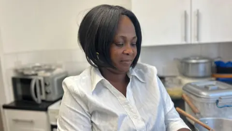 Felicity Afriyie, how has her dark hair cut into a bob and is wearing a white shirt. She sits in a kitchen and is looking down, not at the camera