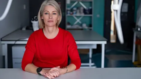 Firecrest scientist Joan Cochrane sits at a table in the forensics lab, wearing a red jumper, a silver bracelet and a thick black watch. She has shoulder length blonde hair. Behind him is a microscope on a table, on the right are some cabinets and on the far right is the main door.