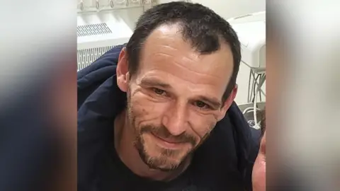 Family A white man with brown hair and facial hair and brown eyes. He wears a navy jumper and smiles at the camera.