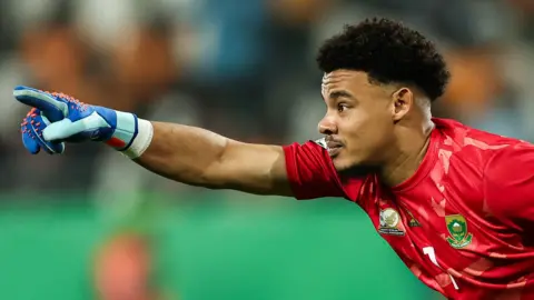 Ronwen Williams, wearing a red South Africa goalkeeping jersey and a blue glove on his right hand, leans forwards into shot from the right-hand side of the image and points with his index finger and arm extended as he looks ahead