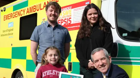 East of England Ambulance NHS Trust Esmai smiles at the camera as she is presented with her award wearing a red hoodie. Neill Moloney crouches next to her who wears a suit with a blue shirt and gold tie. Liam Walker stands behind Esmai wearing a denim coloured shirt while Hollie Kett stands behind Mr Moloney wearing a black jumper and green trousers. They all stand in front of an ambulance.