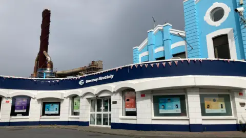 Exterior shot of Guernsey Electricity's headquarters. It is a blue and white building. A large copper chimney is in the background. Grey clouds are in the sky.