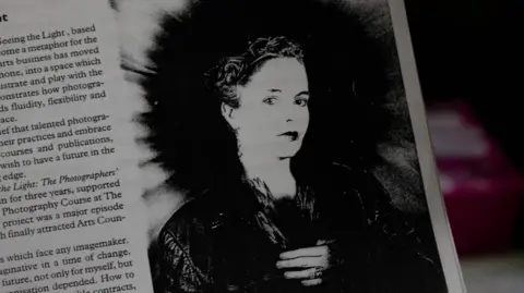 A black and white image of photographer Rhonda Wilson featured in a magazine, with some text to the left of the image. She is wearing a dark jacket and has short hair. 