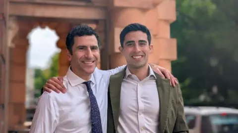 UCH Photo of Prof Hani Marcus and Mr Danyal Khan standing outside. Both have dark brown hair. One is wearing a white shirt and blue and white tie. The other is wearing a light coloured shirt and kaki blazer.