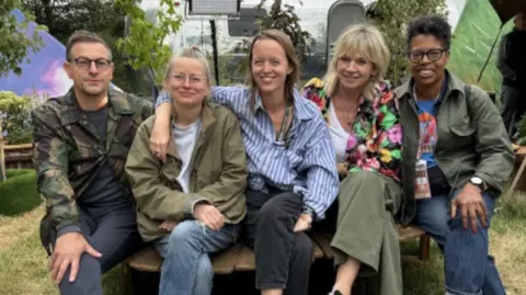 Five people sitting on a wooden chair. Alison Howe has short light-coloured hair and glasses, wearing a green anorak and white T-shirt. Emily Eavis has long brown hair and is wearing a blue and white shirt. There are potted plants behind them.