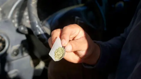 A close-up of a person's hand, holding a roll-up piece of paper with cannabis sprinkled inside. They are sitting at the wheel of a car.