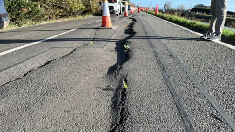 Part of the road with large, wavy cracks in the tarmac and raised sections. Orange and white cones stretch along the middle of the road as cars pass.