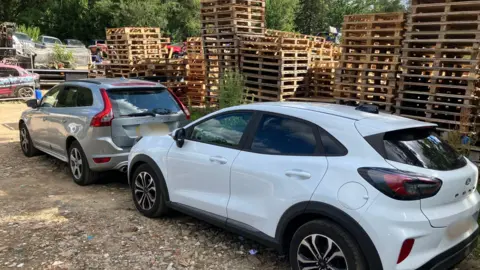 Piers Hopkirk/BBC A grey car and a white car parked next to wooden crates stacked on top of each other.