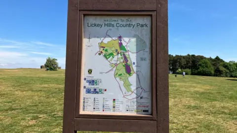 Large grassy area in a park, with trees in the background. A rectangular wooden sign says Lickey Hills Country Park with a map of the park.