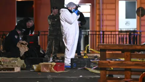 Getty Images, a forensic researcher who uses a white monkey, takes images of the scene, who is covered with boxes and police tape. Behind him, police officers recover evidence.