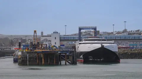 Manx Scenes The Manannan catamaran is a white ship with a dark blue hull, moored at Douglas port, alongside a stone pier with iron pillars going into the water.