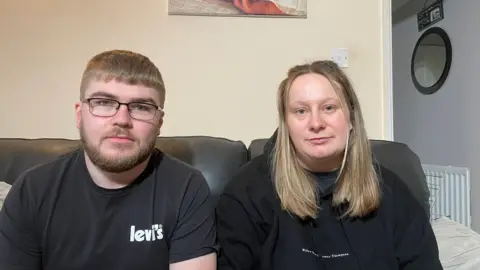 Mr and Mrs Courtney-Thompson, sitting in room with clean walls, staring at the camera