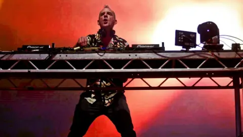 A man in colourful flowery shirt stands on stage behind DJ decks. The stage is lit by pink and red lighting.