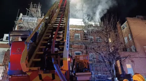 London Fire Brigade A red and silver turntable ladder stretches up and over a converted brick block of flats covered in scaffolding, with smoke rising into the dark sky