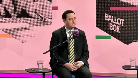 Ben Houchen is sitting in front of a BBC Radio Tees microphone. He is wearing a black suit and yellow-striped tie. The graphic behind him show a ballot box.