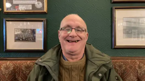 A smiling, bald-headed man wearing glasses and a green coat and an olive coloured jumper.