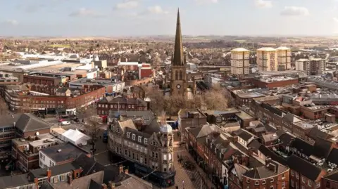 A wide aerial view of Wakefield's city centre surrounded by a dense residential and commercial area. At the centre of the scene stands a tall, pointed church spire made of dark stone, rising prominently above the surrounding buildings. The church beneath the spire is constructed from similar stone and includes a clock face that is visible on the tower.