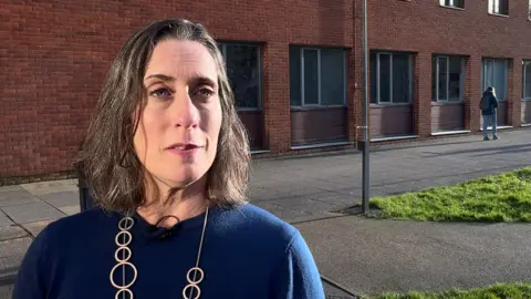 A woman is standing looking towards the camera. She is wearing a blue jumper and a silver ringed necklace. She has brown hair and pink lipstick. There is a path and a red-brick wall behind her. 