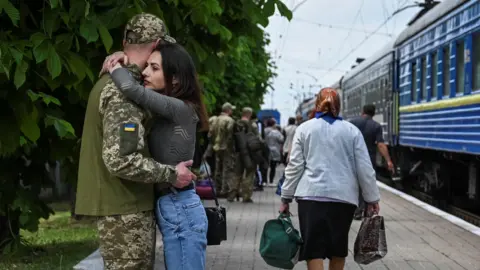 A man in army fatigues hugs a young woman on a train station platform, with a Ukrainian blue and yellow train in the background