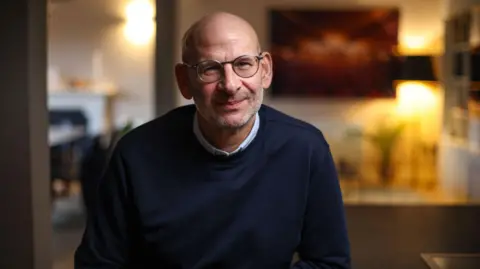 Rogan Productions David Conn, a bald man with glasses, in a dark blue crew-necked jumper and light blue shirt. The background is a domestic room with warm lighting and is slightly out of focus.